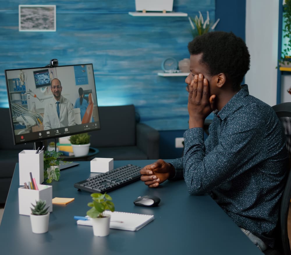 A man is sitting at a computer with his hand on his face. On his screen, you can see he is having a virtual consultation with a clinician.