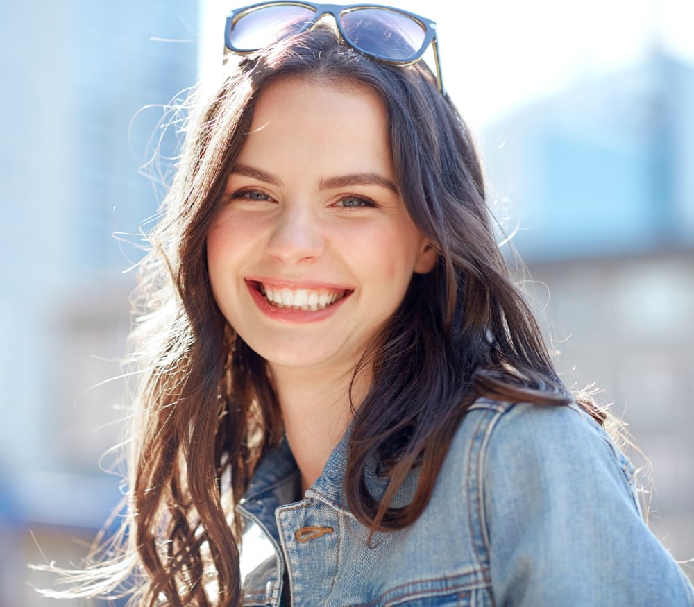 A young girl is smiling. She is wearing a denim jacket and has sunglasses on her head.