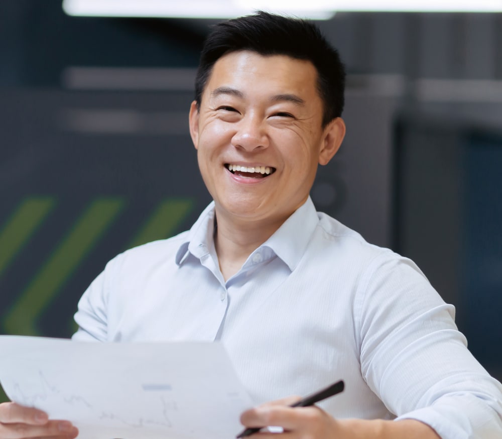 A man is smiling widely. He is holding a document and a pen in his hand.