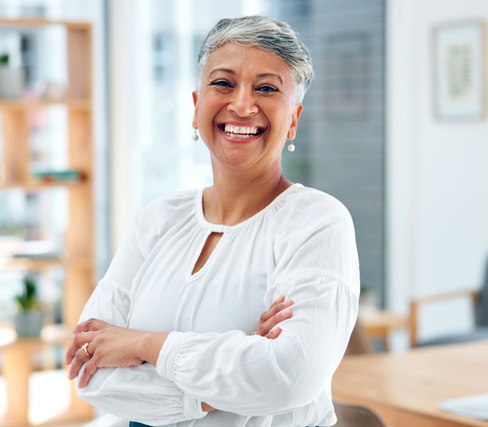 A woman in a white blouse is smiling. She has her arms crossed.