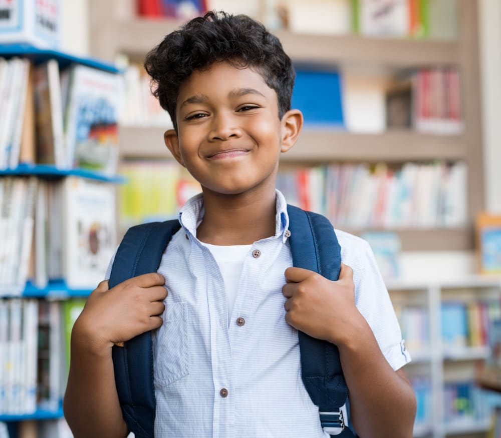 A young boy with curly hair is smiling and holding on to his backpack straps.